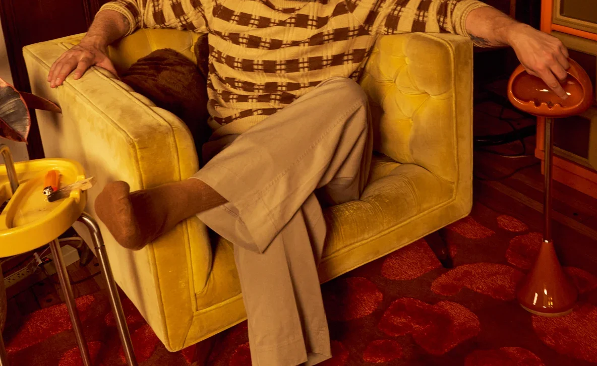 Someone sunk into a yellow velvet armchair on a red rug, a yellow side table ashtray on one side and an orange standing ashtray on the other