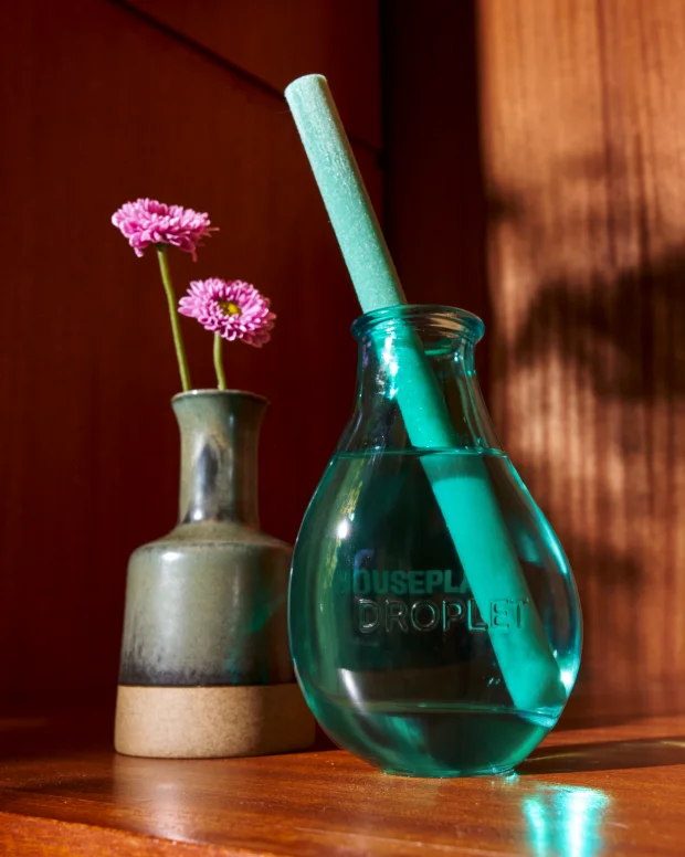 A teal glass diffuser with a single reed beside a small vase of pink flowers