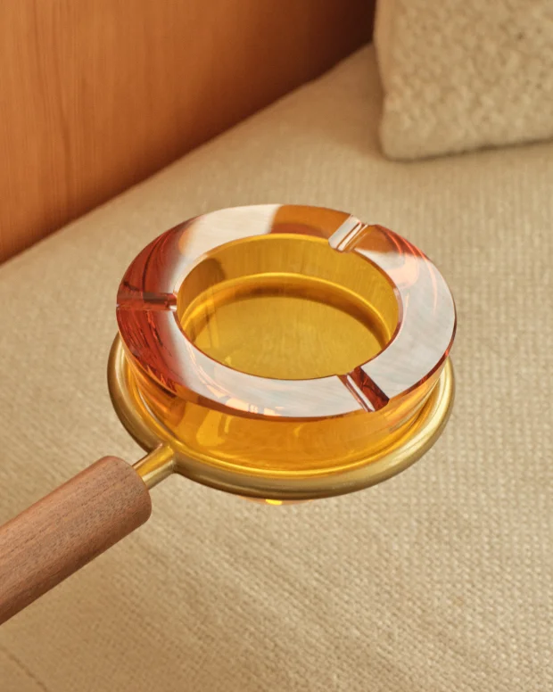 An amber glass ashtray on a brass arm with a wooden handle, against a cream sofa
