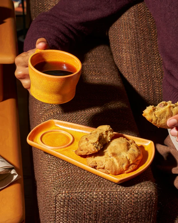 A hand holding an orange ceramic cup above a matching snack tray on the arm of a sofa