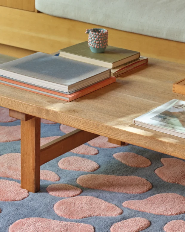 A blue rug with pink gloop shapes under a wooden coffee table stacked with books