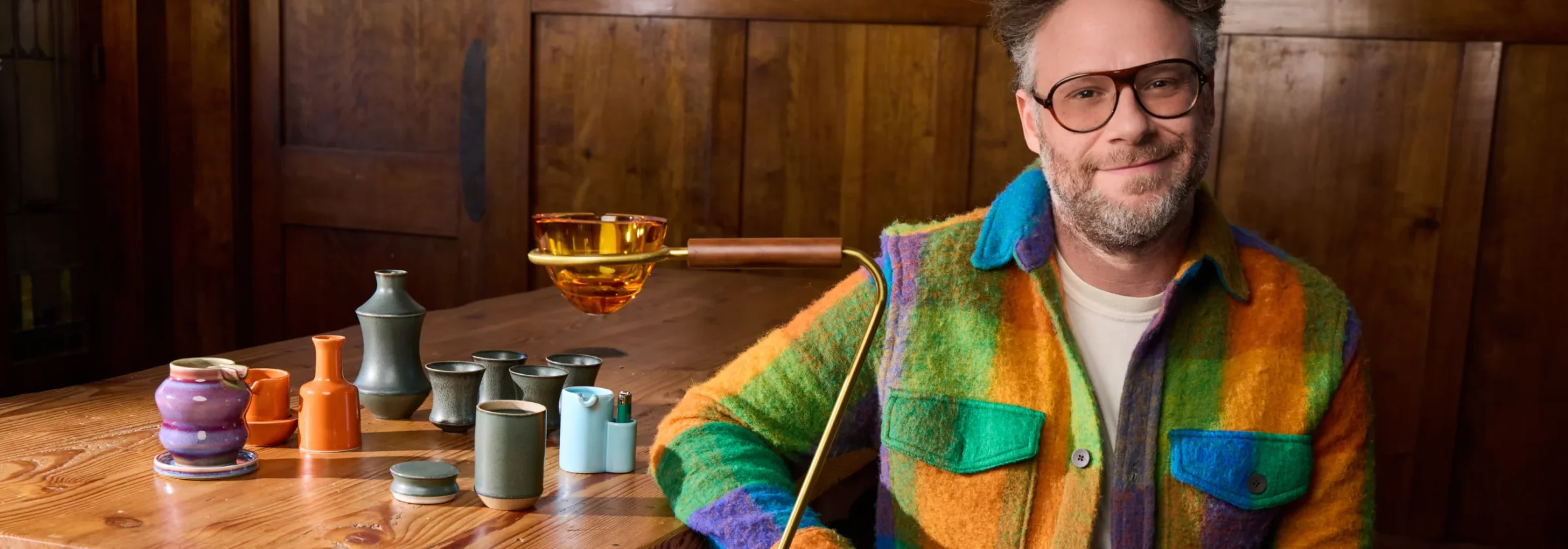 Seth Rogen at a wooden table with a line-up of ceramic ashtrays, vases and an amber standing ashtray