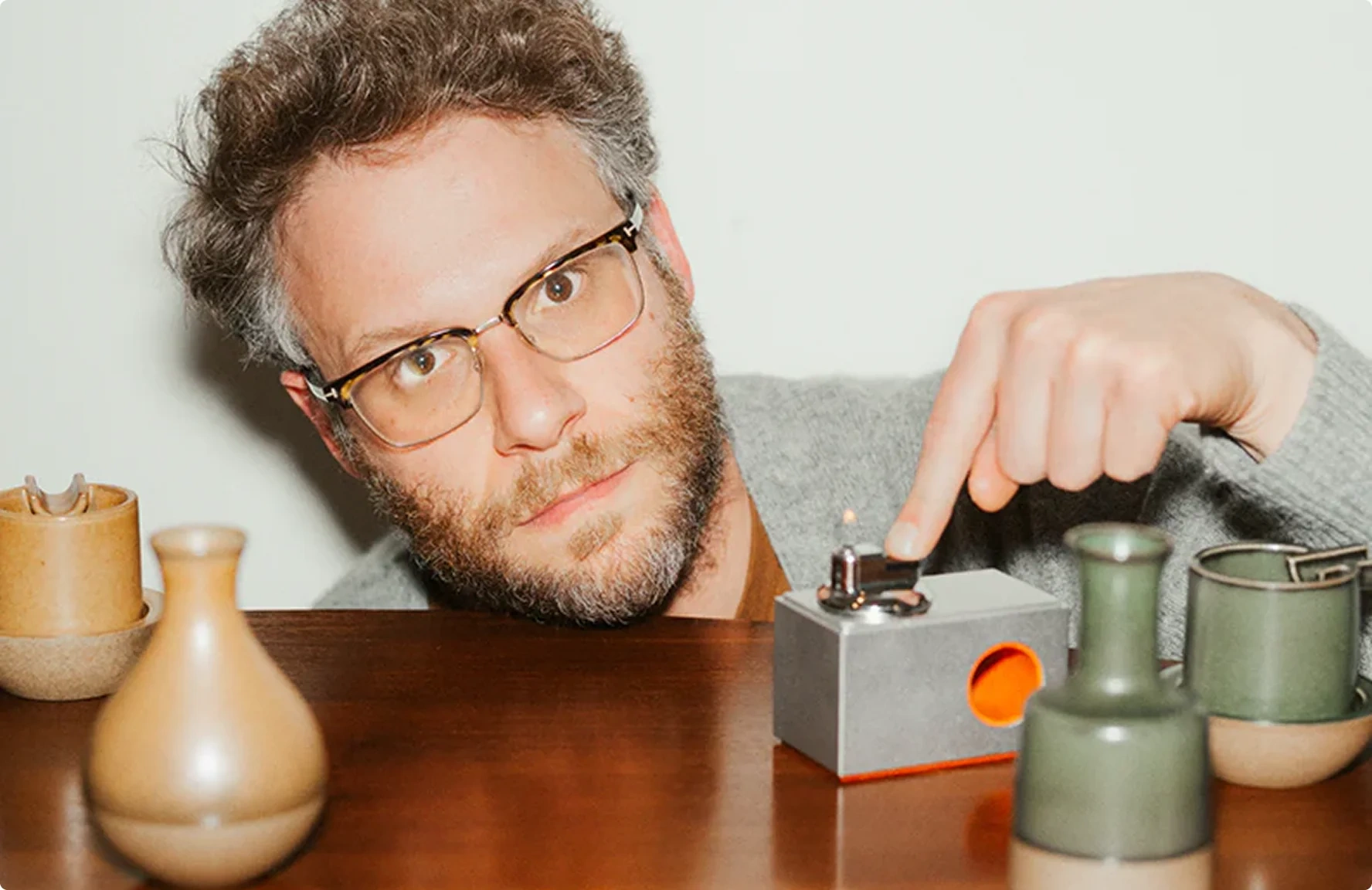 Seth Rogen leaning on a table, pressing a finger onto a small grey gadget beside ceramic vases